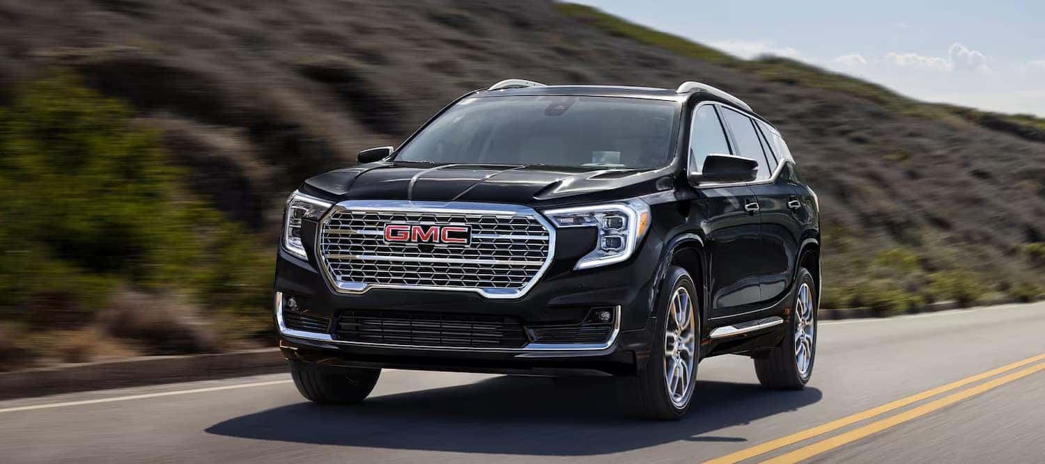 Black GMC SUV driving on a road flanked by hills under a clear blue sky.