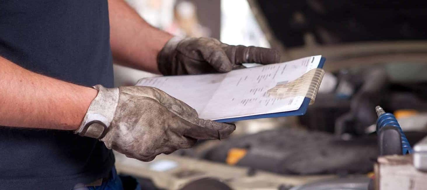 Mechanic holding a clipboard in a garage, wearing dirty gloves, with a car engine in the background.