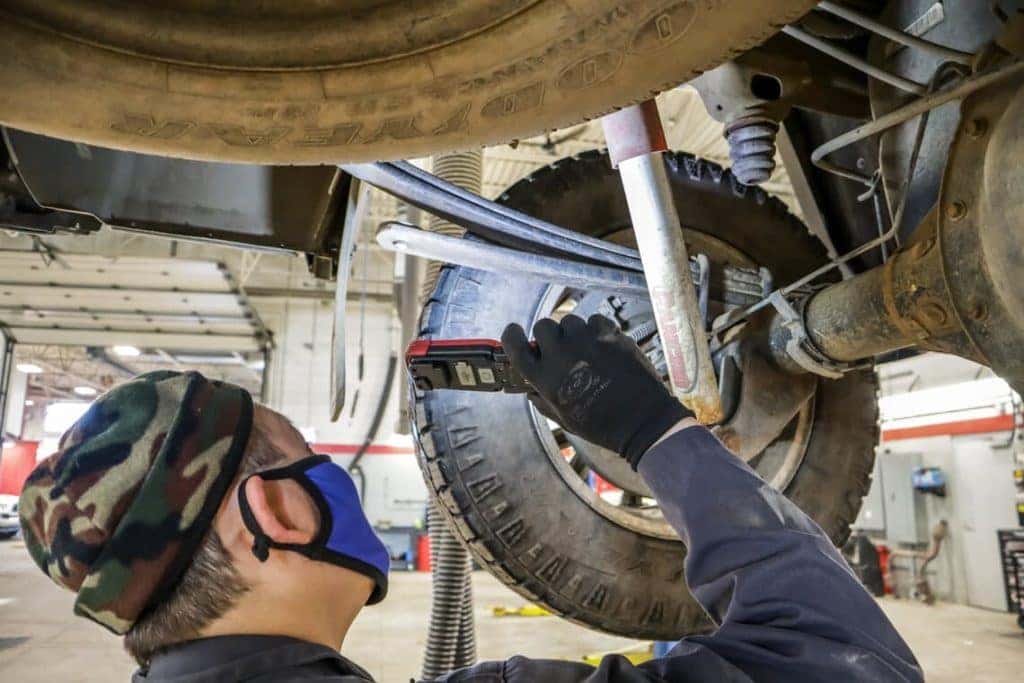 Mechanic inspecting a vehicle's suspension and wheel in a garage setting, wearing a mask and gloves.