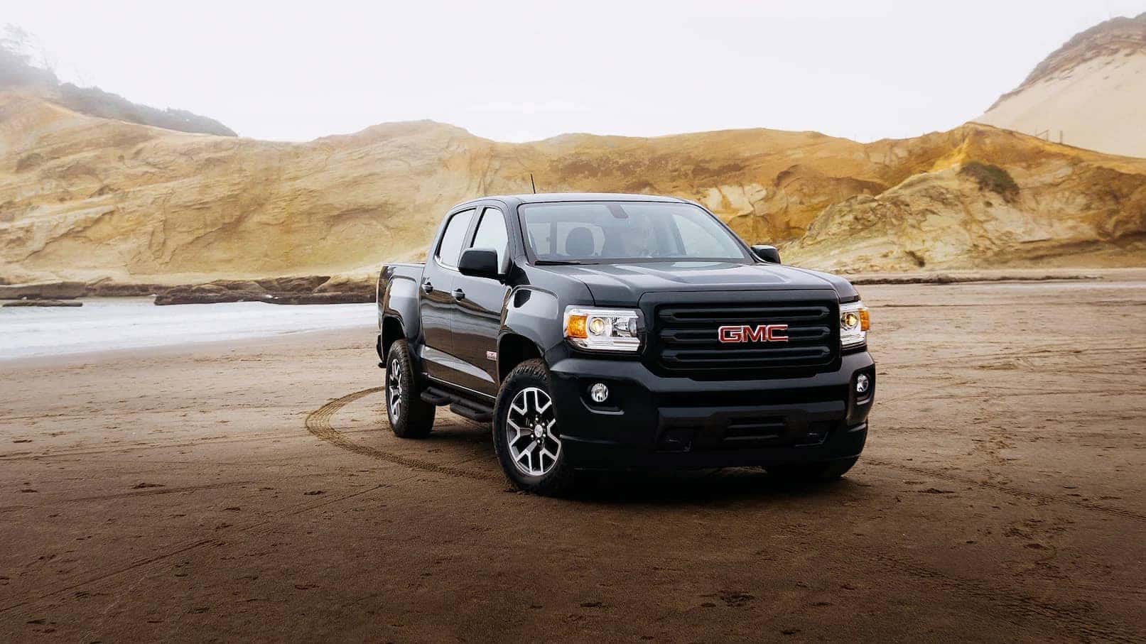 Black GMC pickup truck parked on a sandy beach, with rocky cliffs in the background.