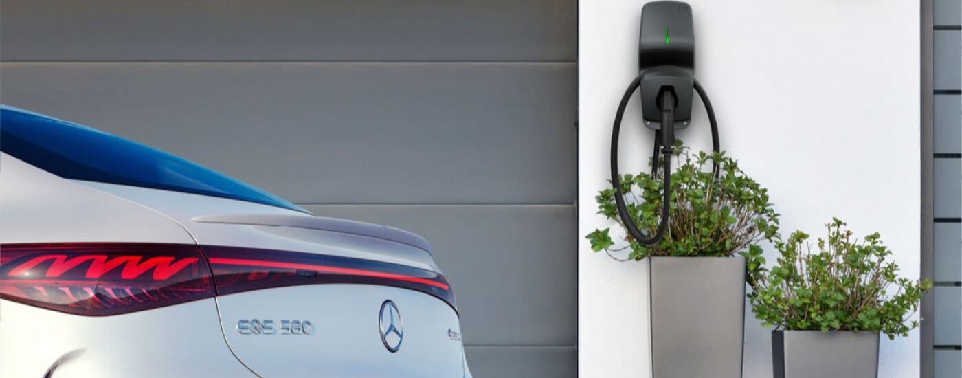 Rear view of a Mercedes EQS 580 charging at a wall-mounted EV charger, with potted plants nearby.
