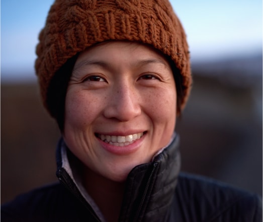 Person smiling warmly, wearing a brown knit hat and black jacket, outdoors at twilight.