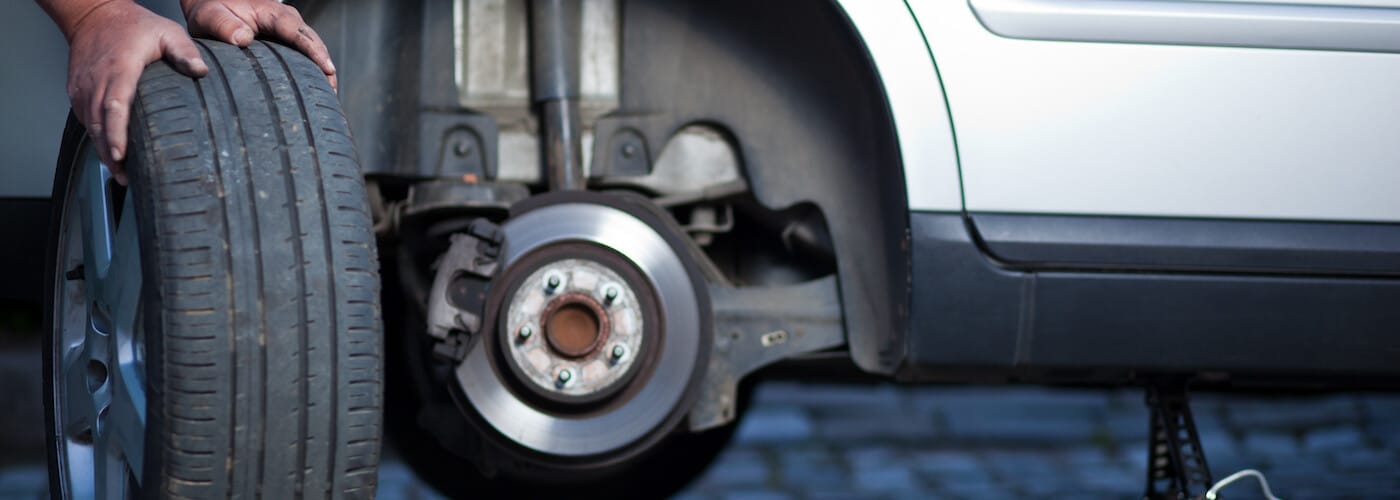 Person changing a car tire, with hands holding the tire near the vehicle's exposed brake rotor.