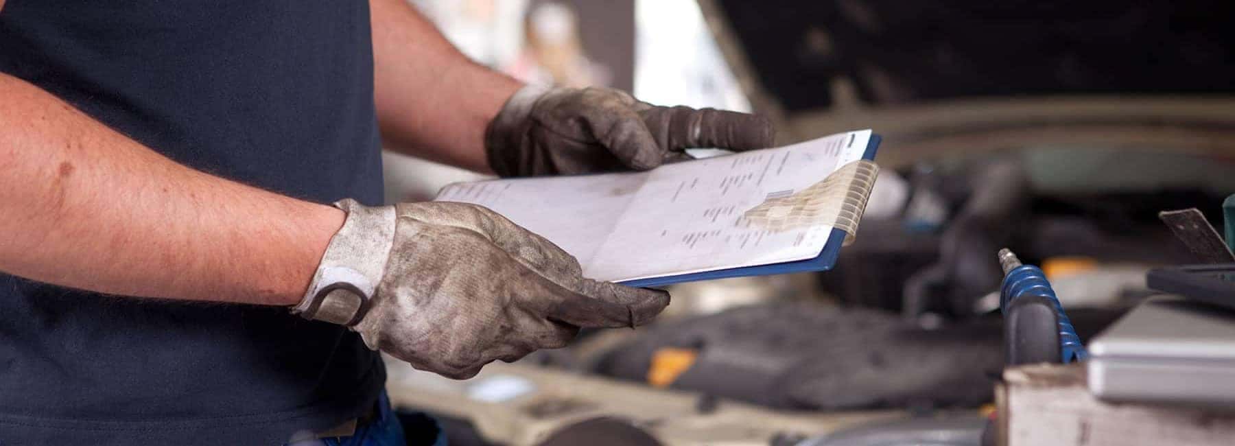 Mechanic with gloves holding a clipboard, inspecting a car engine in a workshop.