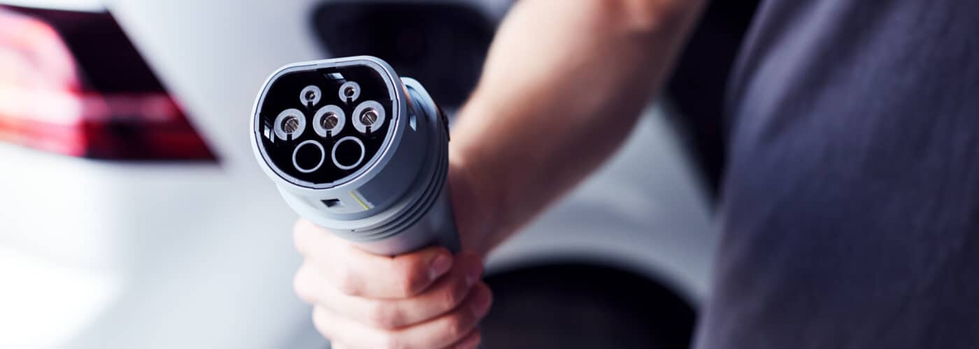 Person holding an electric vehicle charging plug, car in the background.